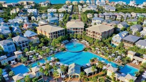 An aerial view of a resort with multiple buildings, a central swimming pool, and surrounding palm trees.