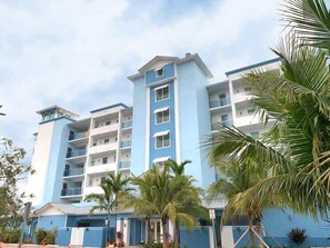 A multi-story hotel with a blue and white exterior, palm trees in the foreground, and a clear sky. - Waterfront Captain's Stateroom: Views, Pool & Hot Tub! (Merritt Island)