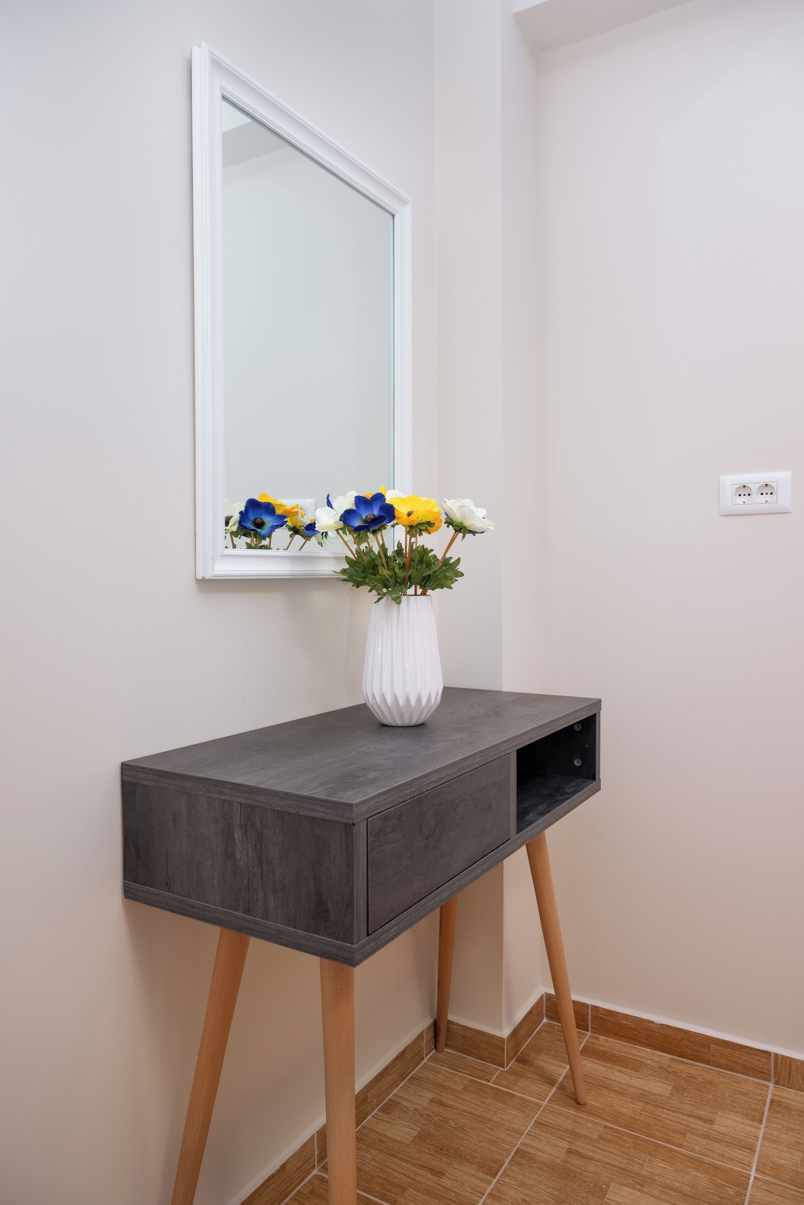 Une console grise surmontée d’un vase blanc rempli de fleurs, avec un miroir au-dessus et une prise électrique sur le mur.