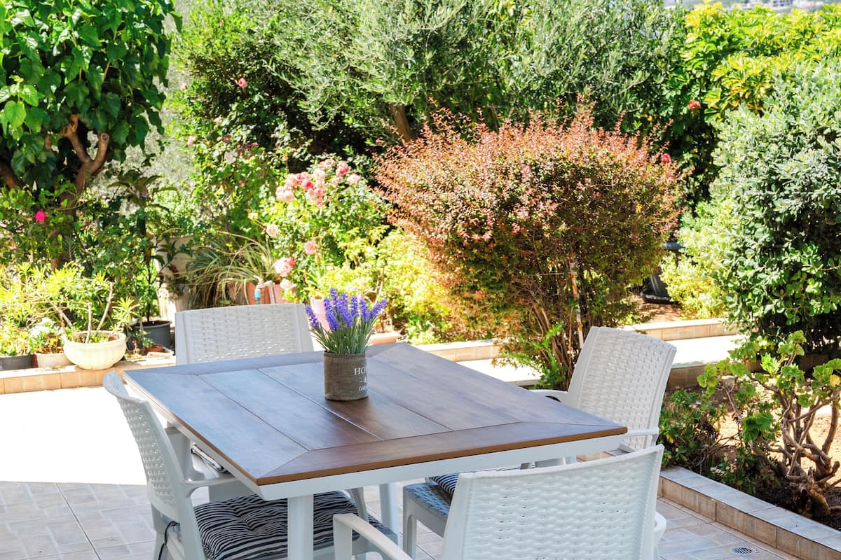 An outdoor patio with a wooden table and white wicker chairs, surrounded by potted plants and trees.