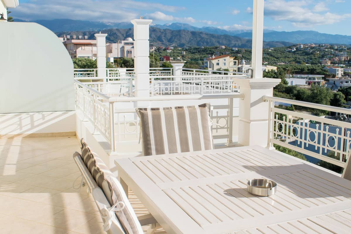 A terrace with a white table and chairs, a metal bowl, and a view of a town and mountains in the background.