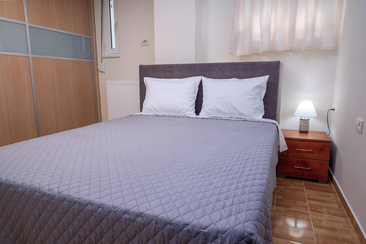 A neatly made bed with a quilted cover, a bedside table with a lamp, and a wooden wardrobe.