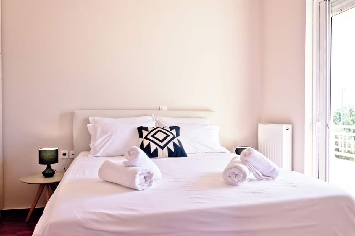 A neatly made bed with white linens and two rolled towels, a bedside table with a lamp, and a window with a view of greenery.