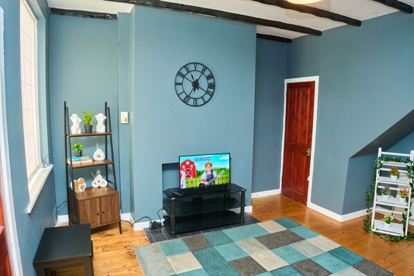 A living room with a television displaying a children's show, a black entertainment unit, a wooden cabinet, a clock, and a staircase. - 2dbed Close to Etihad, Coop Live, Manchester (Manchester)