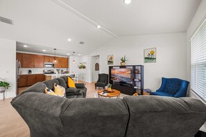 A modern living room with a grey sofa, a coffee table, and a TV. There is a kitchen in the background with wooden cabinets and a microwave.