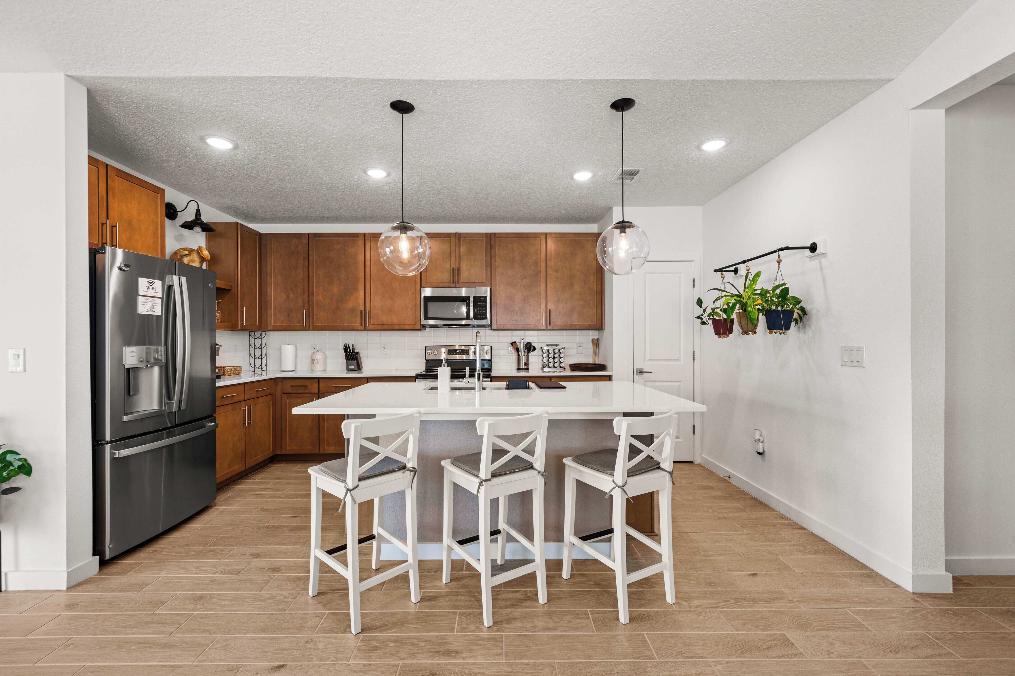 A modern kitchen with wooden cabinets, stainless steel appliances, a white island with bar stools, and pendant lighting.