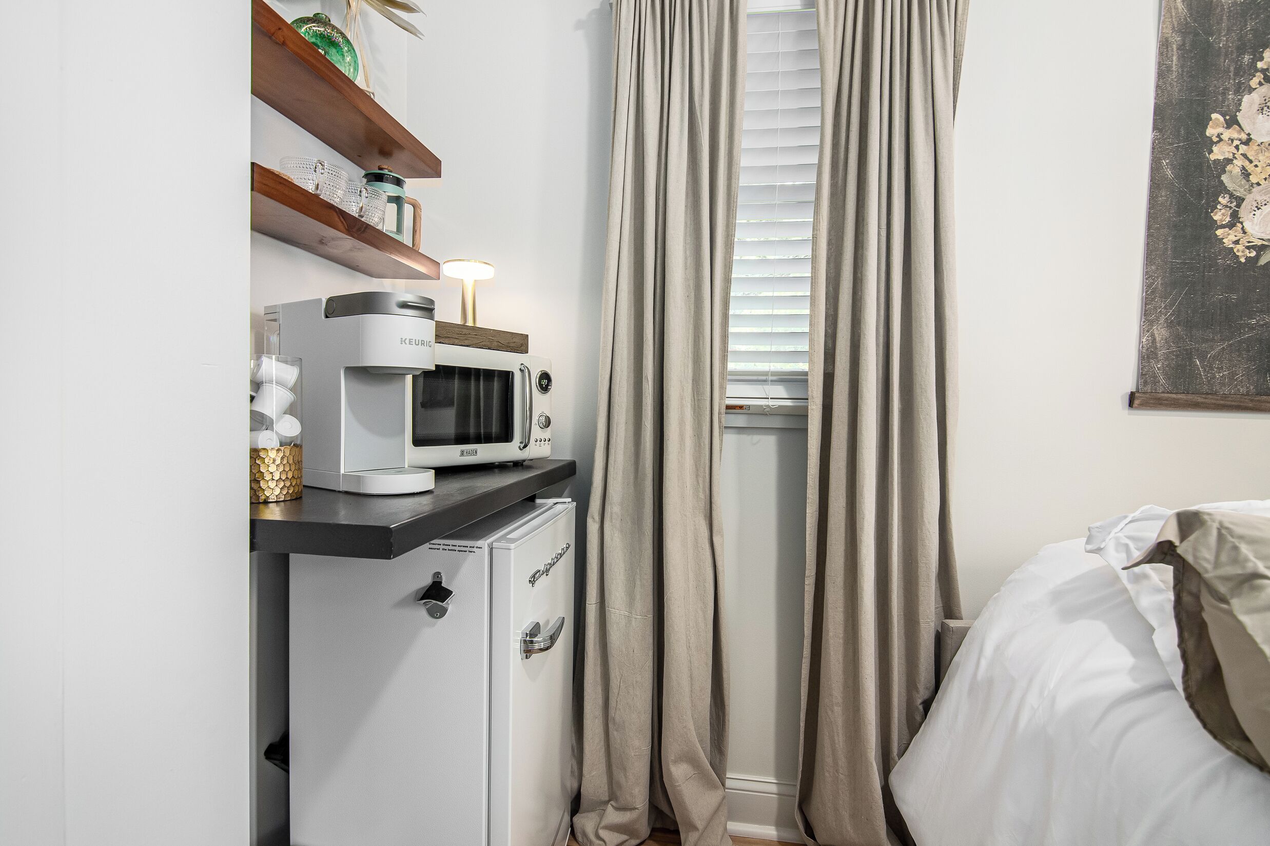 Un dormitorio compacto con cama, una kitchenette pequeña con microondas y un ventanal con cortinas.