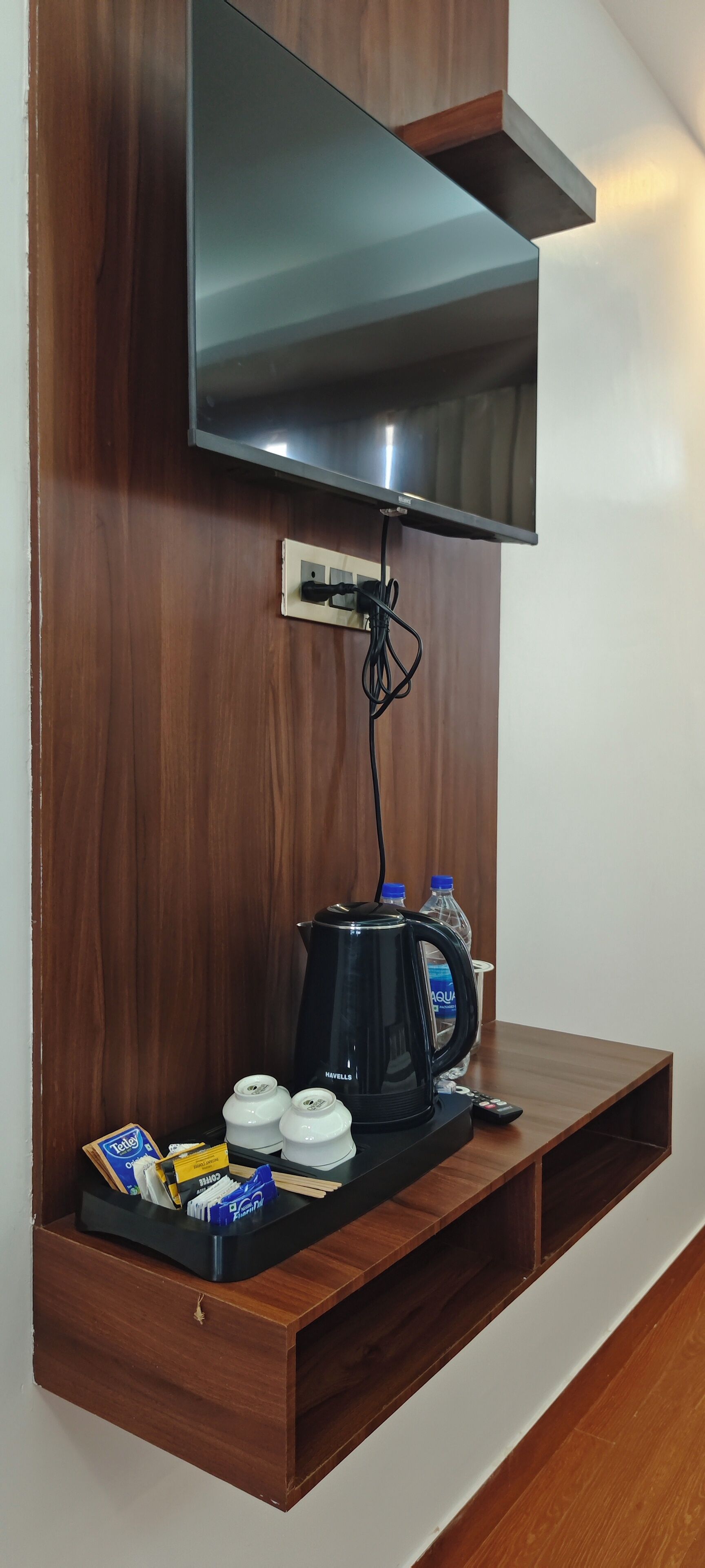A wooden wall with a flat-screen TV mounted above a wooden shelf. On the shelf, there is a black coffee maker, two white teapots, a remote control, and a water bottle.
