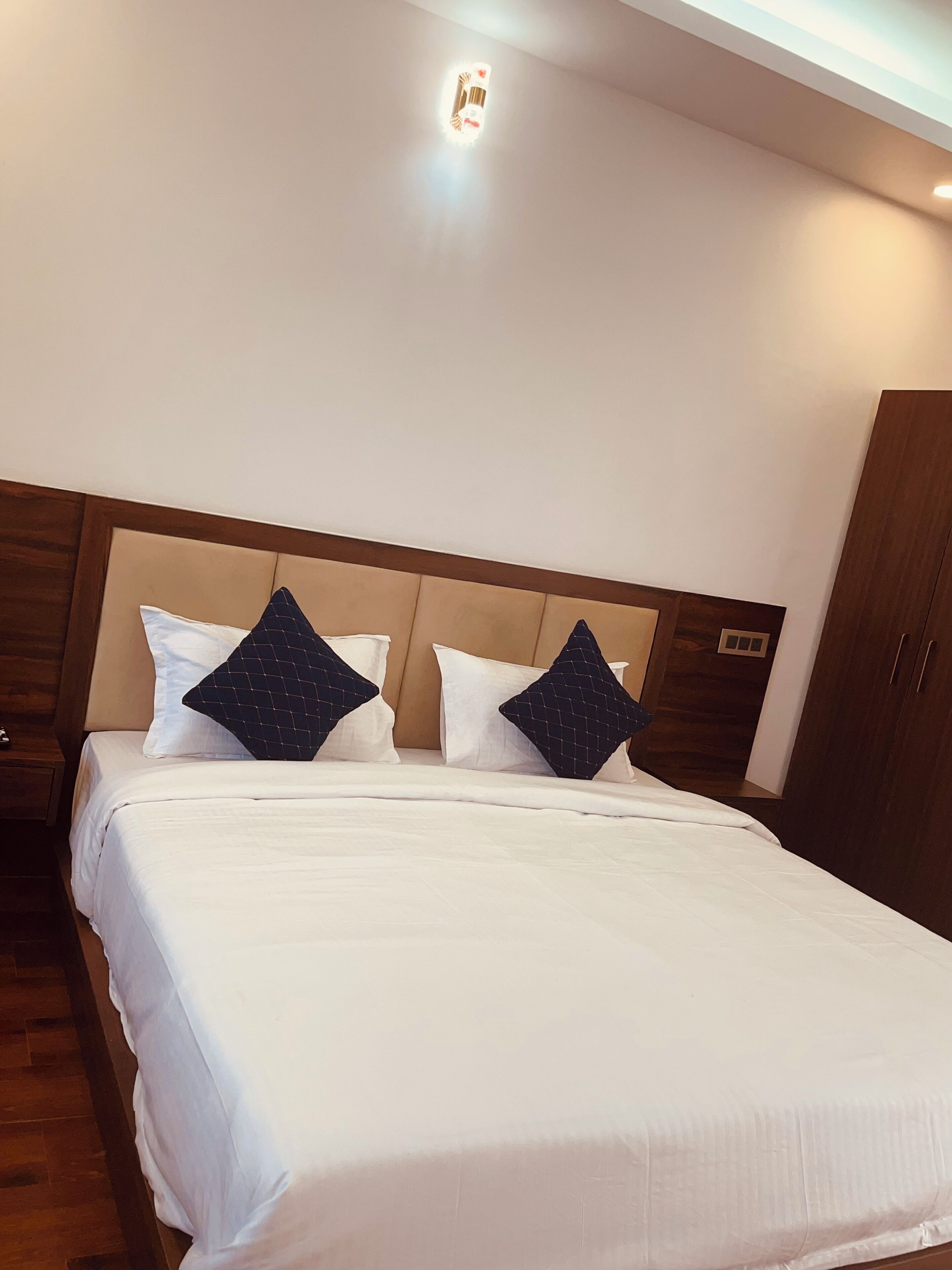 A neatly made bed with white linens and two dark pillows, a wooden headboard, and a nightstand with a lamp.