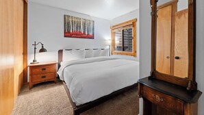 A bedroom with a bed, a wooden nightstand, a desk, a mirror, and a window with shutters.