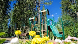 A playground with a slide and climbing structures, surrounded by trees and flowers.
