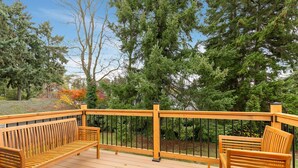 A wooden deck with a bench and a table, surrounded by trees and a metal railing. - Renovated Home | Dogs OK | Large Deck (Seattle)