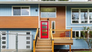 A two-story house with a red door, wooden steps, and a small porch with a bench. - Renovated Home | Dogs OK | Large Deck (Seattle)