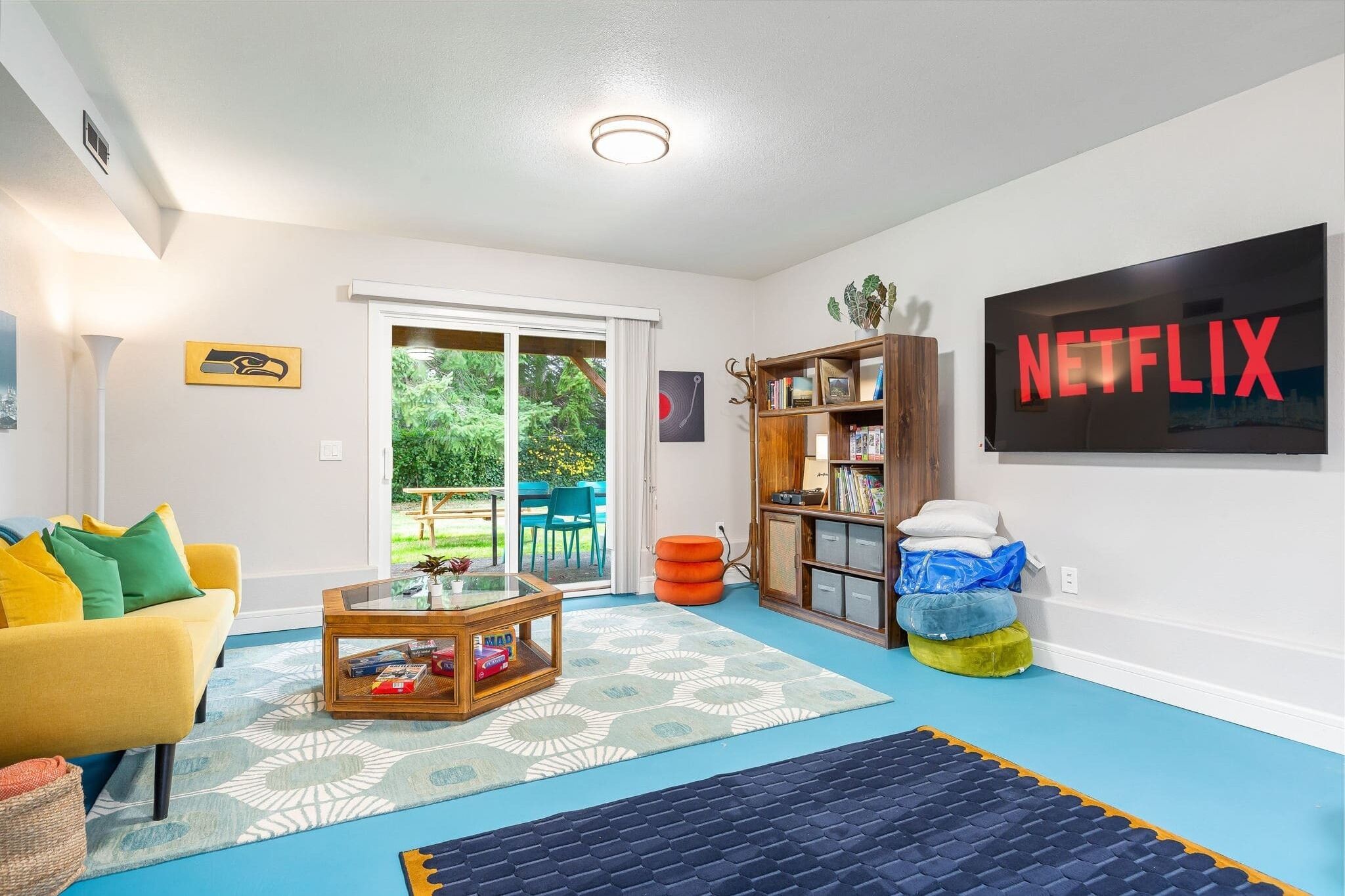 A living room with a yellow sofa, a coffee table, a bookshelf, and a large TV screen displaying the Netflix logo.