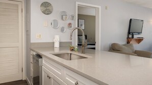 A modern kitchen with a large island, stainless steel sink, and a built-in oven. There are multiple wall-mounted mirrors and a flat-screen TV on a wooden shelf. - Summit Unit 1103 2 bed ground floor, pet friendly condo, ski-in/ski-out access (Granby)