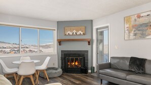 A living room with a fireplace, a gray sofa, a dining table with chairs, and a view of a snowy landscape through large windows. - Summit Unit 1103 2 bed ground floor, pet friendly condo, ski-in/ski-out access (Granby)