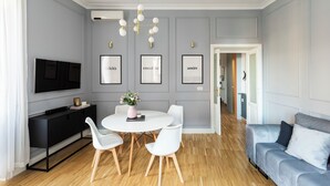 A modern living room with a grey sofa, a white dining table with chairs, a black entertainment unit, and a TV mounted on the wall. - Charming Home near San Giovanni (Rome)