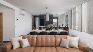A modern living room with a large brown leather sofa, a dining area with a long table, and a kitchen in the background. - Penthouse Adela (Vir)