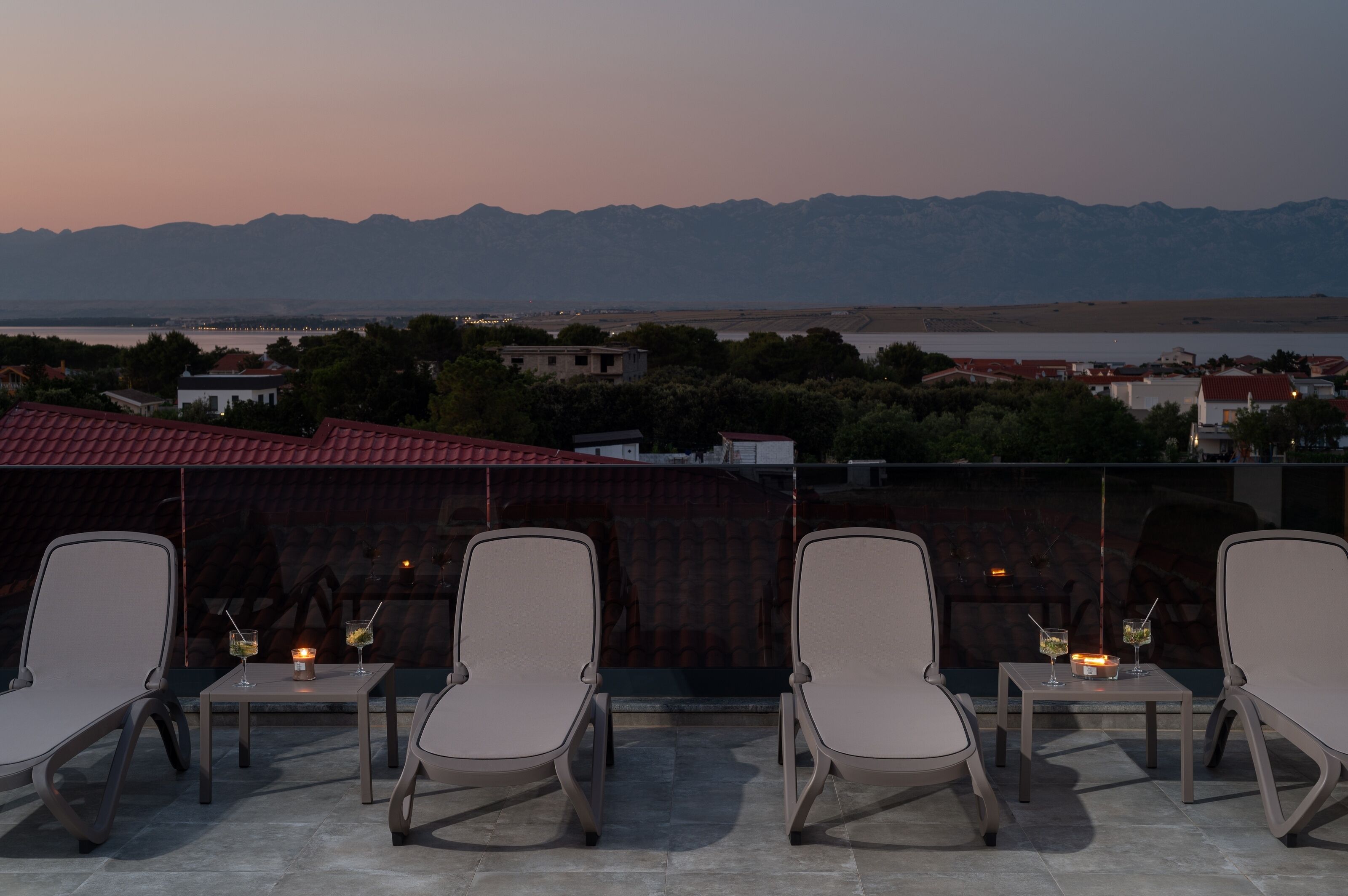 A terrace with four lounge chairs and two tables, each with a drink and a candle, overlooking a mountainous landscape and a body of water.