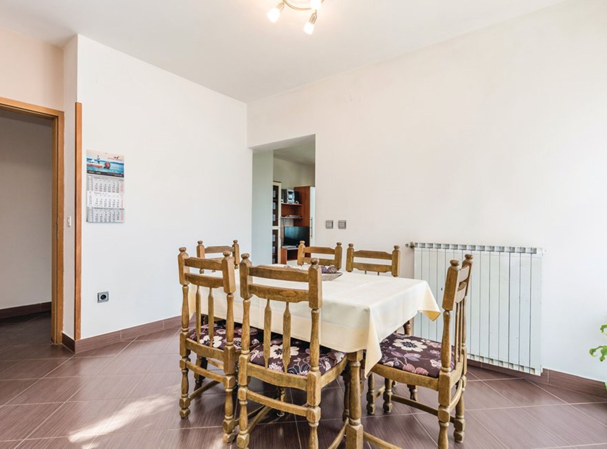 A dining area with a table and four chairs, a radiator, and a calendar on the wall.