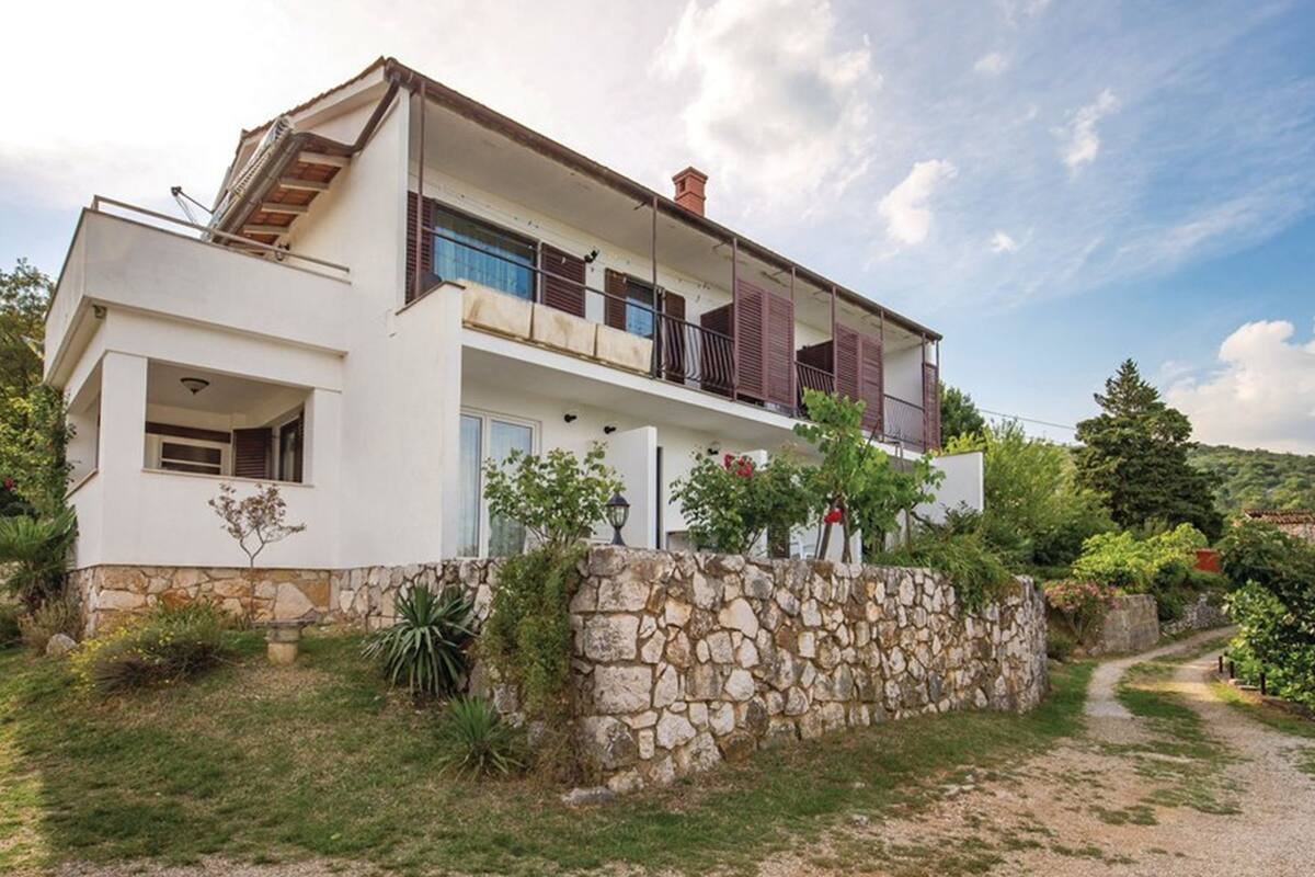 Une maison moderne à deux étages, dotée de fondations en pierre, d’un balcon avec des volets en bois et d’un jardin où poussent différentes espèces de plantes.