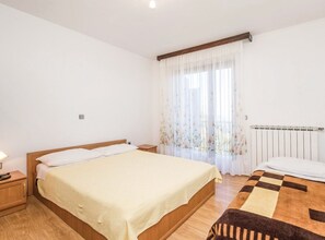 Un dormitorio con cama, mesita de noche, radiador y ventana con cortinas traslúcidas.