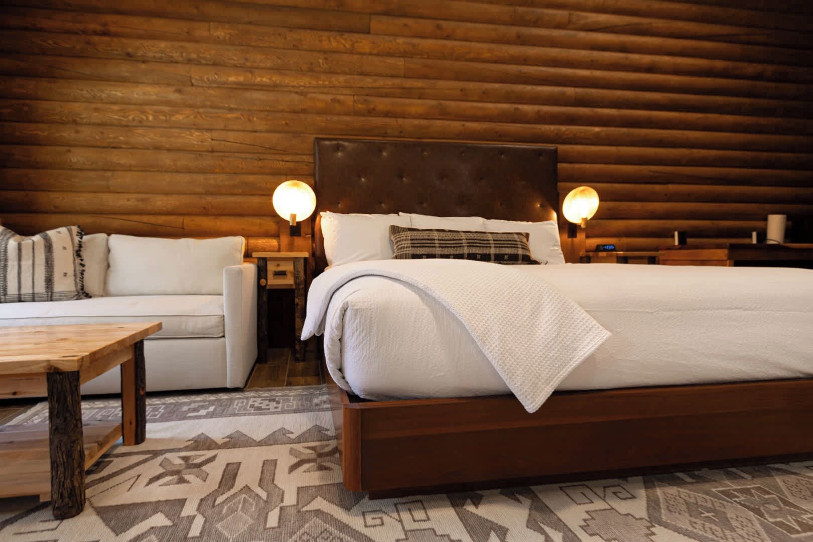 A cozy log cabin-style bedroom with a large bed, a wooden headboard, two bedside lamps, a wooden nightstand, a white sofa, and a patterned rug.