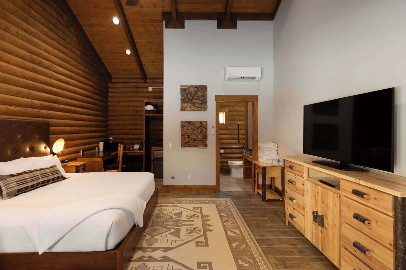 A log cabin-style bedroom with a large bed, wooden headboard, a flat-screen TV, and a wooden dresser.