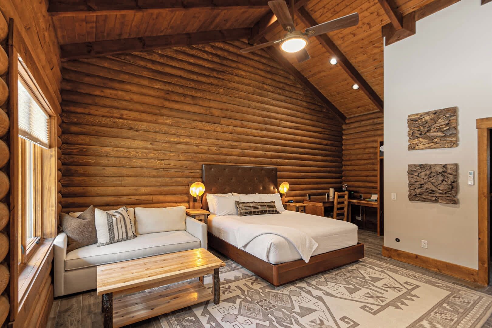 A log cabin bedroom with a bed, a sofa, a wooden bench, and a patterned rug.