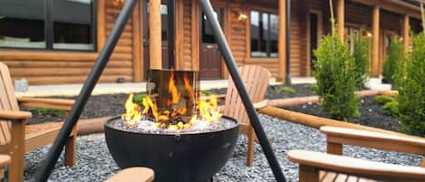 A fire pit with chairs around it, set in a gravel area with a wooden building in the background.