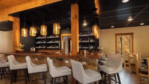 A bar area with wooden beams, hanging pendant lights, and a stone wall. There are bar stools and a bar counter with glassware.