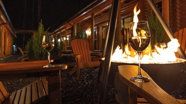 An outdoor seating area with wooden tables and chairs, a fire pit, and glasses of wine.