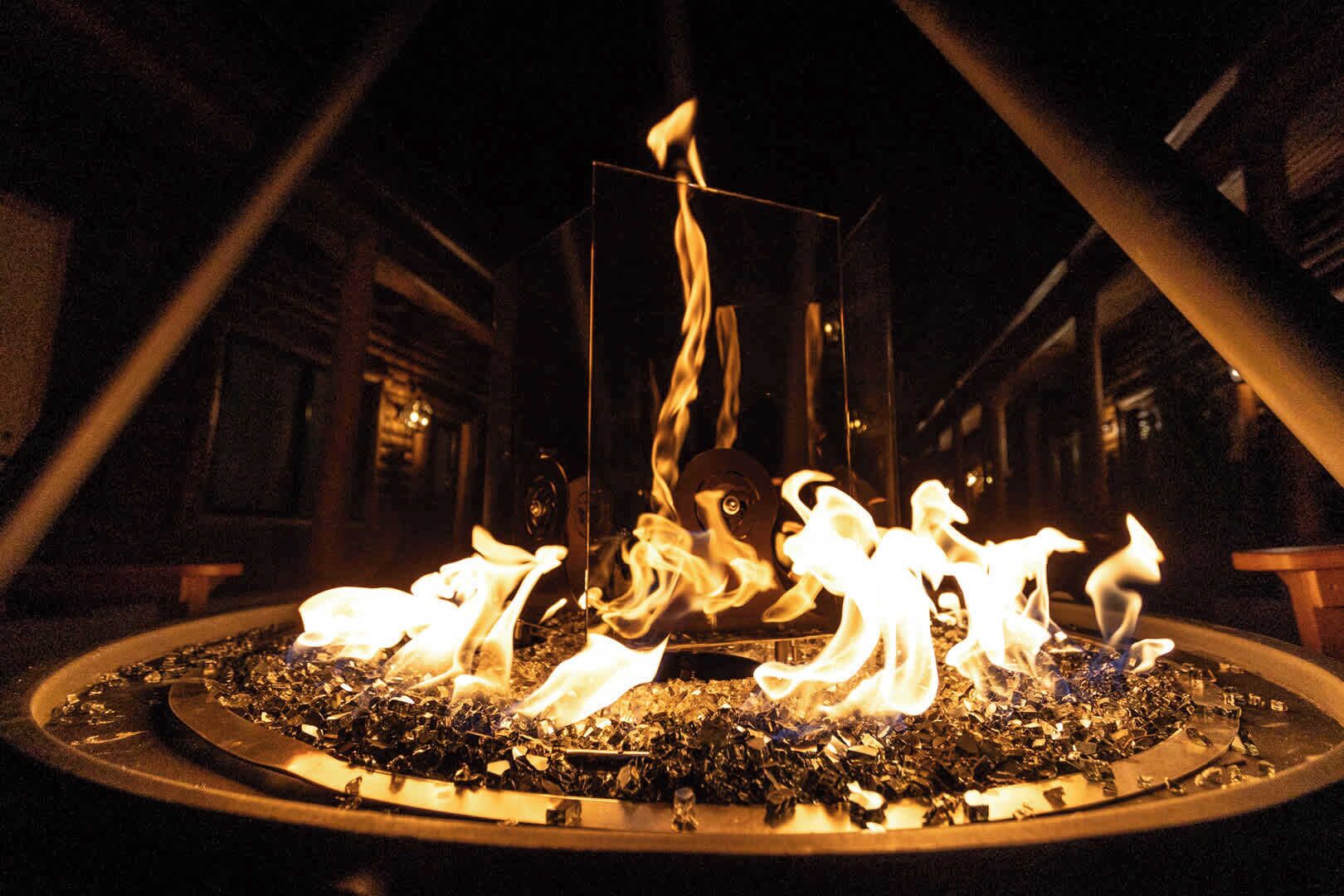 Un foyer dont les flammes s’élèvent dans un récipient métallique circulaire, le tout intégré dans un décor industriel sombre.