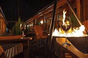 An outdoor seating area with wooden tables and chairs, a fire pit, and glasses of wine.