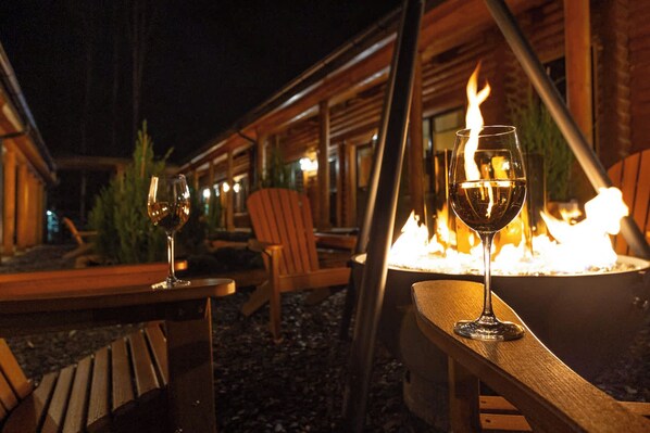 An outdoor seating area with wooden tables and chairs, a fire pit, and glasses of wine.