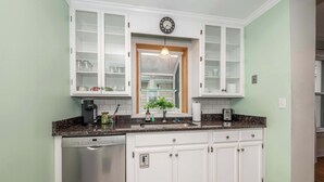 Une cuisine avec des meubles blancs, un lave-vaisselle, une horloge et une fenêtre avec une plante.