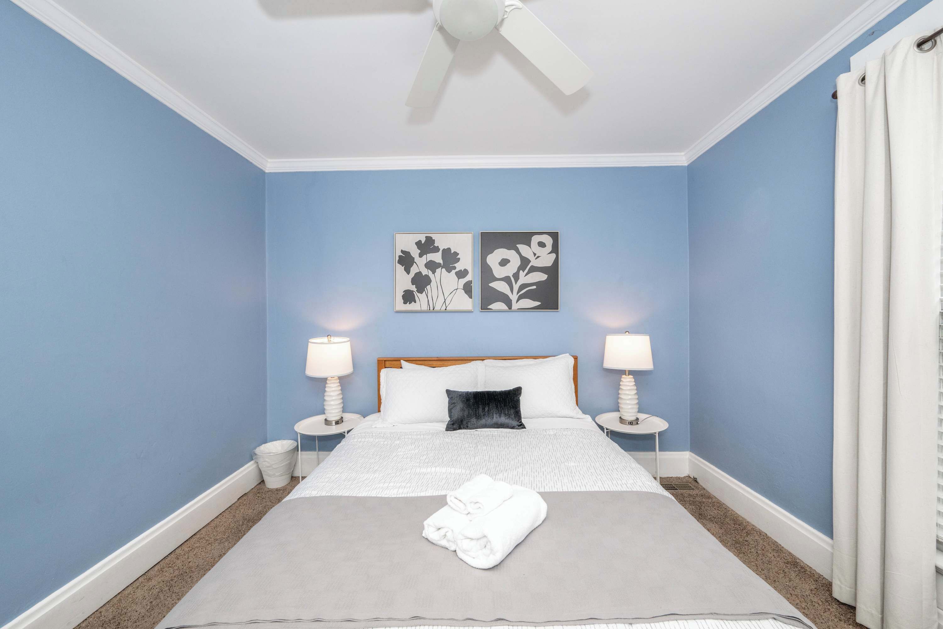 Une chambre à coucher avec un lit, deux tables de chevet avec des lampes, un ventilateur de plafond et des œuvres d’art accrochées au mur.