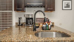 A kitchen with granite countertops, a stainless steel sink, a coffee maker, and a coffee and tea sign.