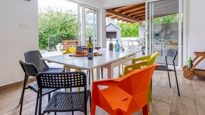 A dining area with a table set for four, surrounded by colorful chairs, and large windows offering a view of greenery outside. - Serene Rural Villa With Pool, Sun Deck, and Children's Play Area (Omis)