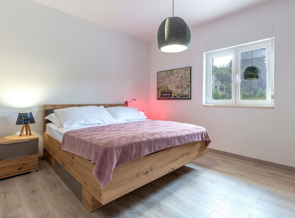 Une chambre avec un lit en bois, une table de chevet avec une lampe, une œuvre d’art représentant une carte accrochée au mur, et une fenêtre donnant sur un paysage verdoyant.