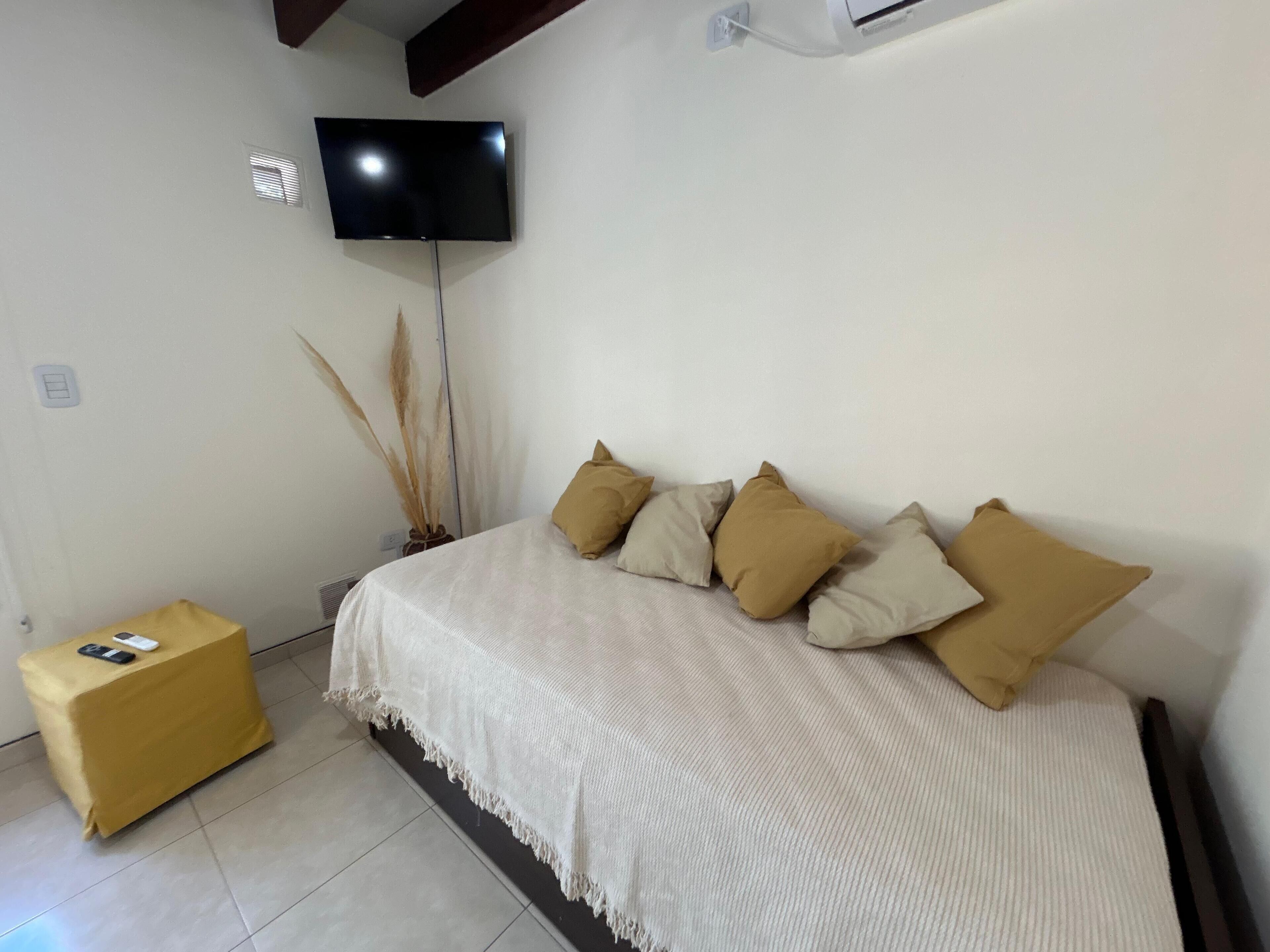 Un dormitorio con una cama, un televisor montado en la pared, una mesita pequeña y una lámpara de noche.