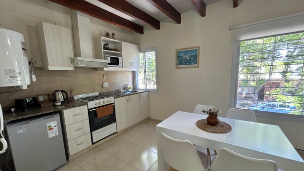 Cocina compacta con gabinetes blancos, microondas integrado, estufa a gas y refrigerador pequeño. Hay una mesa de comedor con dos sillas y un jarrón de flores.
