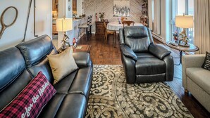 Ein Wohnzimmer mit Sofa, Sessel und einem gemusterten Teppich. Es gibt einen Essbereich mit Tisch und Stühlen, und im Hintergrund eine Küche.