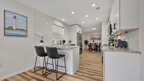 A modern kitchen with white cabinetry, a central island with bar stools, a dining area with a view of the ocean, and a painting of a lighthouse. - The Oak Hideaway | Pet Friendly 3Bd! (St. Augustine)
