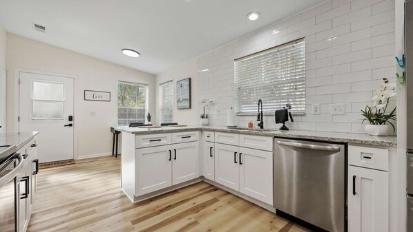 A modern kitchen with white cabinetry, a stainless steel dishwasher, and a window with blinds.