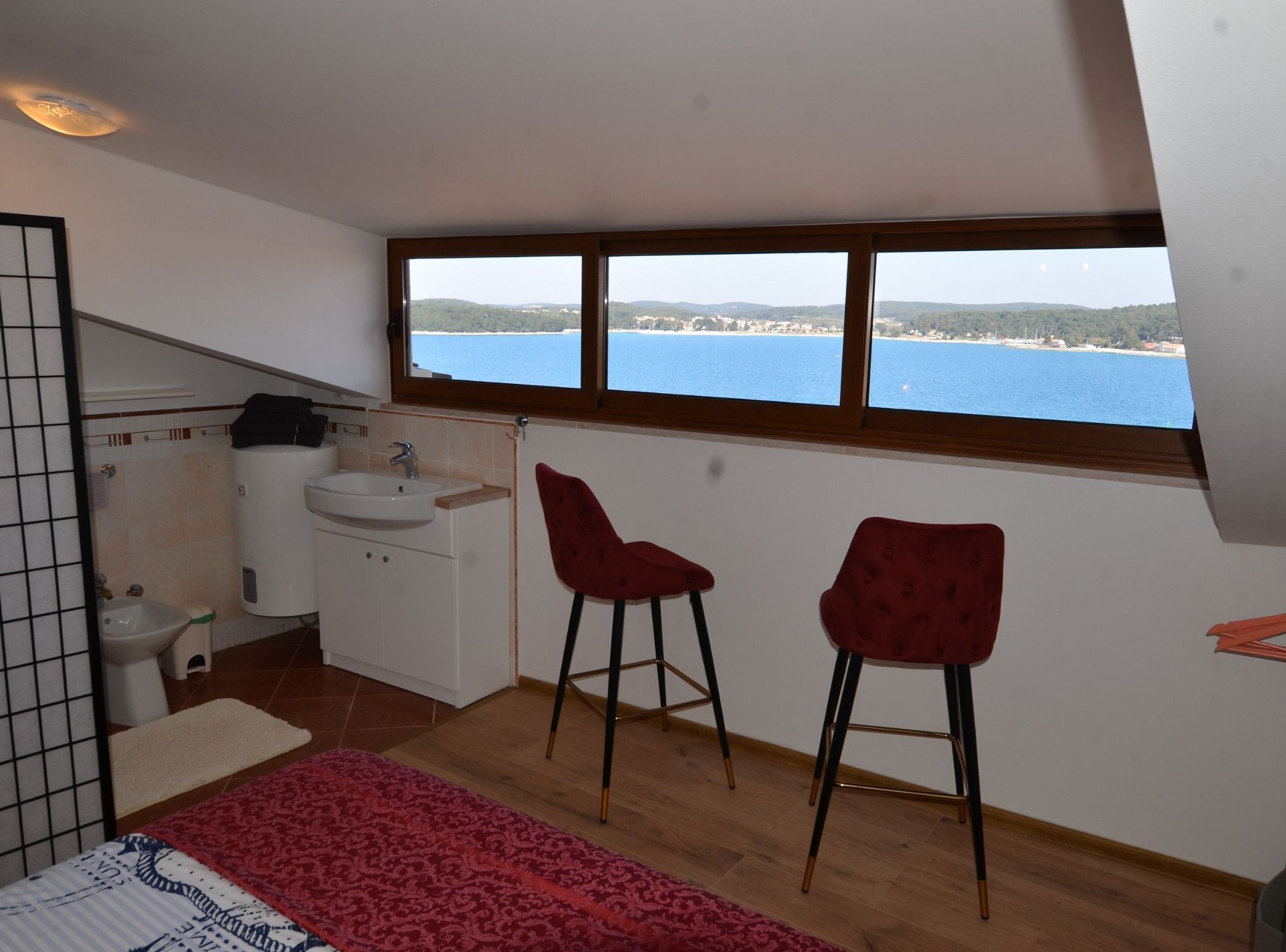 Una habitación pequeña con vista al mar, un lavamanos, dos sillas rojas y una cama con un tapete de diseño rojo y blanco.