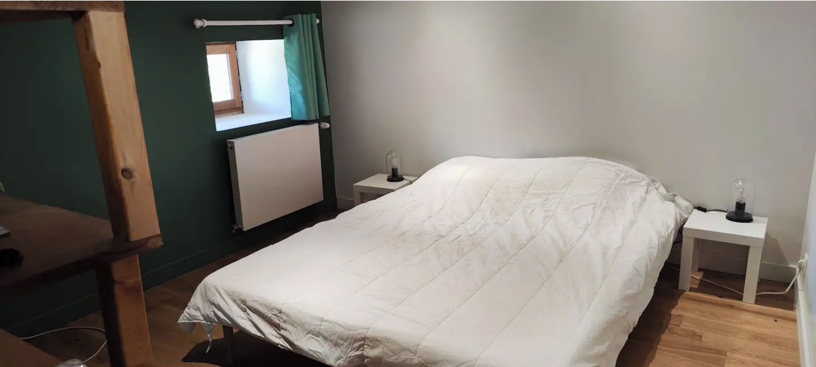 A bedroom with a bed, two bedside tables, a window with curtains, a radiator, and a wooden desk.