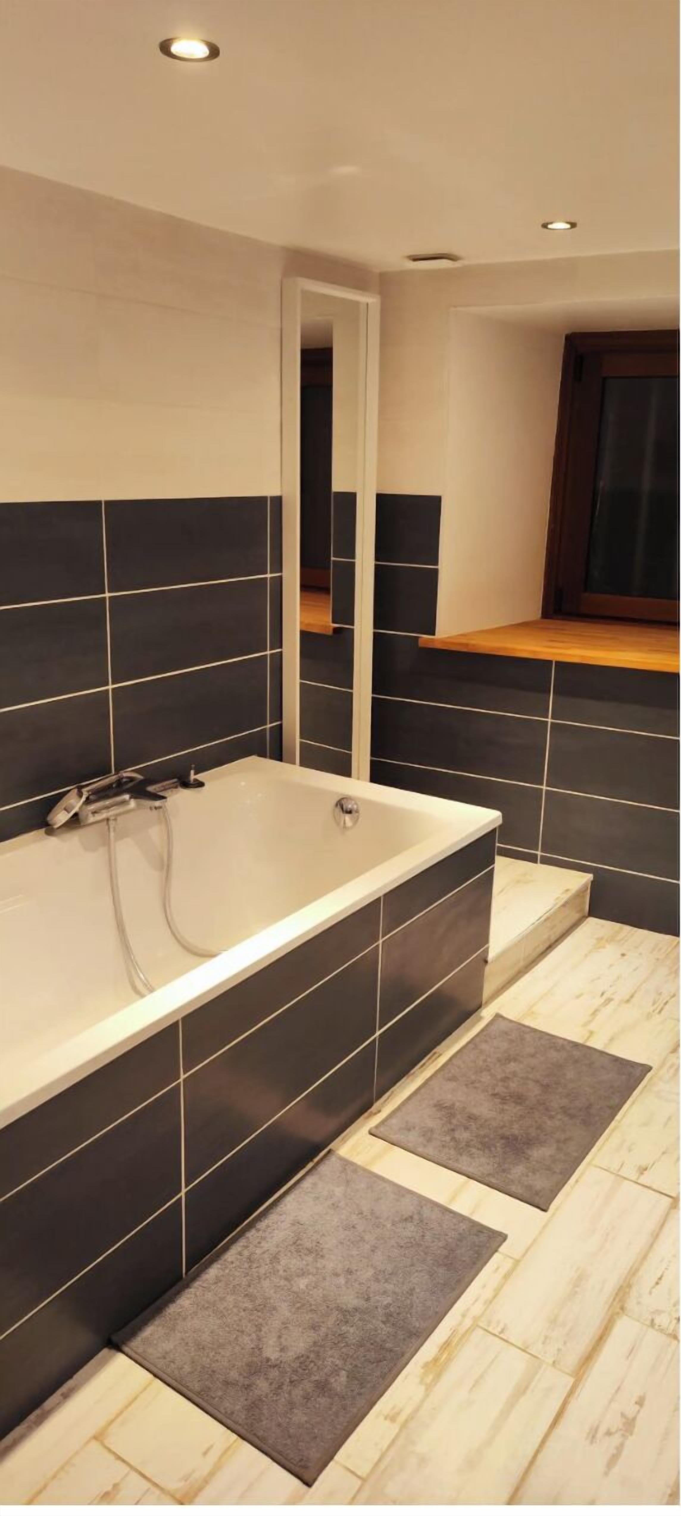 Une salle de bain moderne avec une baignoire sur pieds, des murs recouverts de carreaux foncés et un meuble-lavabo en bois.
