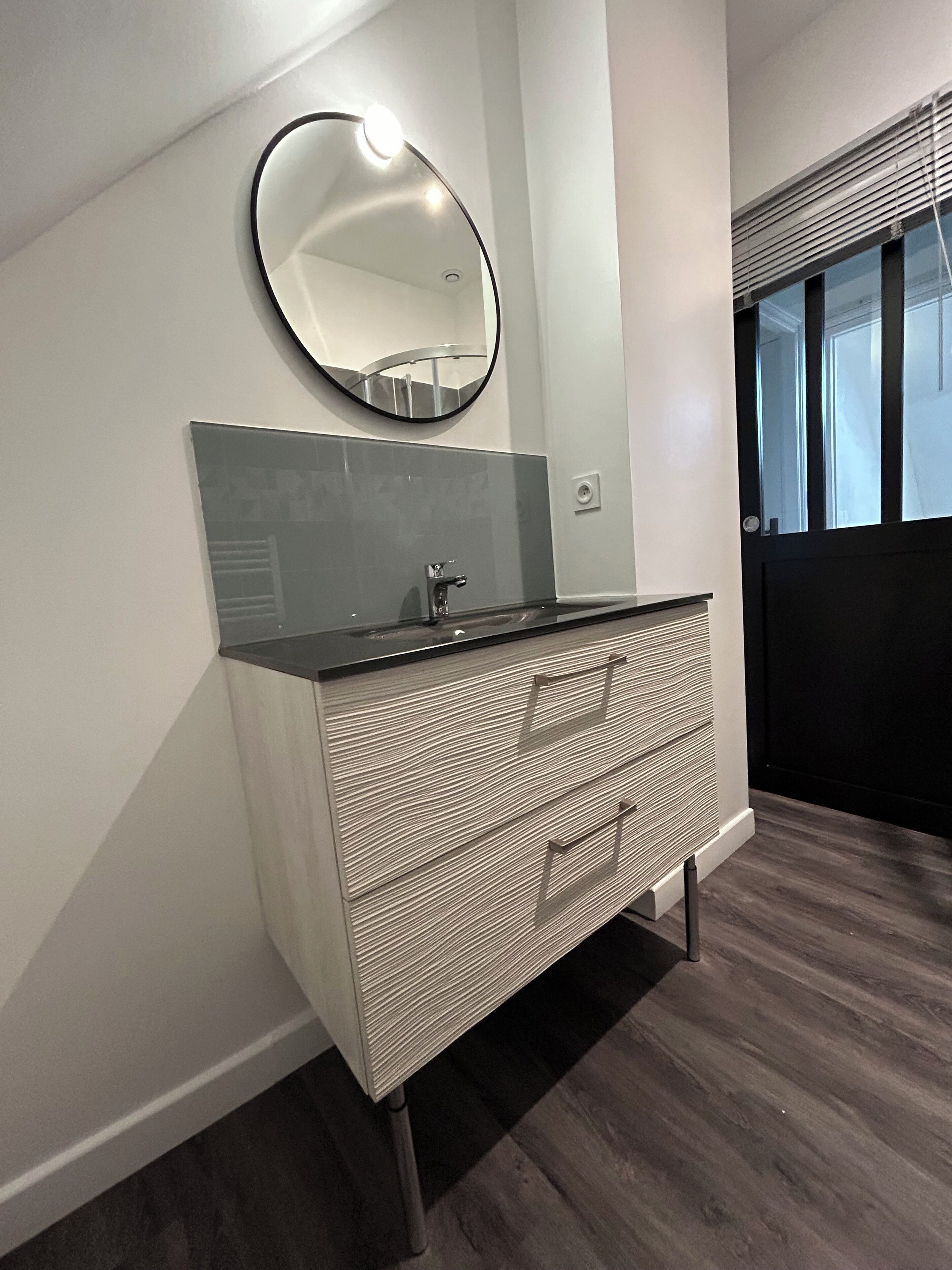 A modern bathroom with a round mirror, a dark countertop, and a patterned drawer unit.