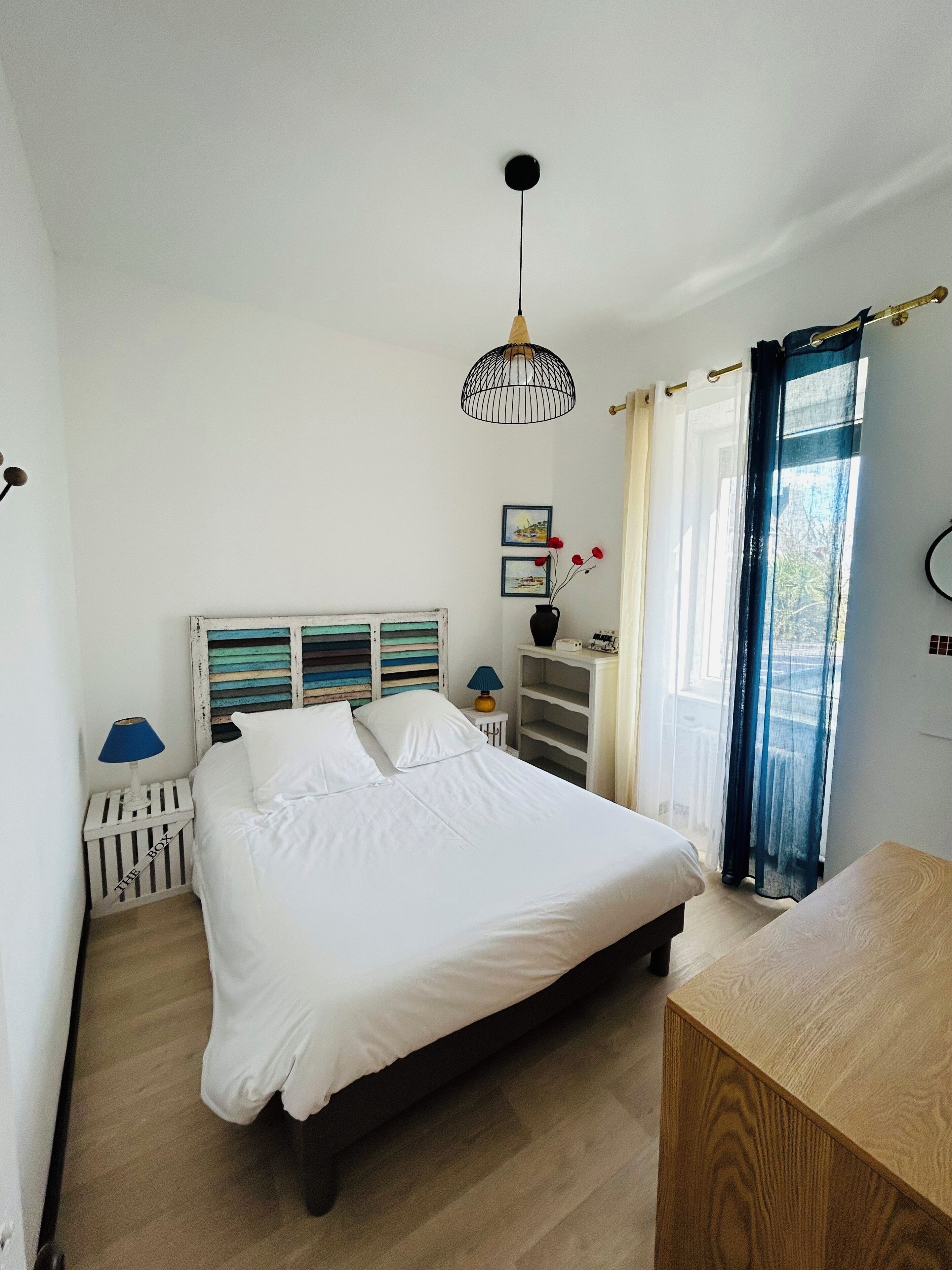 Une chambre à coucher comprenant un lit, une table de chevet, une lampe, une étagère avec des objets décoratifs et une fenêtre avec des rideaux.